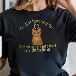 Pode incluir: Camiseta preta com um gato laranja e o texto "I'm Not Ignoring You, I've simply reached my data limit" em letras douradas. O gato tem olhos verdes e está centralizado na camiseta.