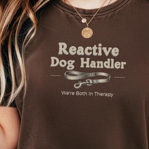 May include: A dark brown t-shirt featuring the text Reactive Dog Handler in a light beige font, with a graphic of a dog leash underneath and the additional text We're Both In Therapy. The word ESPRESSO is printed in the bottom left corner.