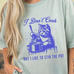 May include: A light sage green t-shirt featuring a blue graphic of a cat stirring a pot with a spoon. The text above the cat reads "I Don't Cook" and the text below the cat reads "BUT I LIKE TO STIR THE POT" in a bold, blue, sans-serif font.
