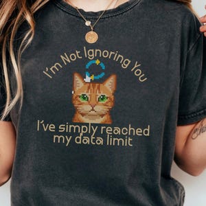 Pode incluir: Camiseta cinza escuro com o texto "I'm Not Ignoring You, I've simply reached my data limit." A camiseta apresenta um gato laranja pixelado com olhos verdes e um gráfico azul e amarelo acima da cabeça.