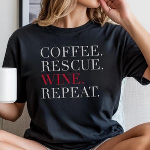 May include: Black t-shirt featuring the text "COFFEE. RESCUE. WINE. REPEAT." in white and red. A casual top, perfect for everyday wear, with a simple, bold statement.