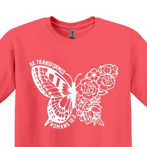 Può includere: T-shirt in seta corallo con un design a farfalla bianca e il testo "Be Transformed" e "Romani 12:2".