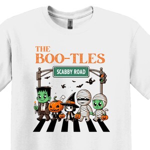 Op de afbeelding: Wit T-shirt met een afbeelding van een groep cartoonmonsters die een straat oversteken. De tekst "The Boo-Tles" en "Scabby Road" staat op het bord boven de straat.