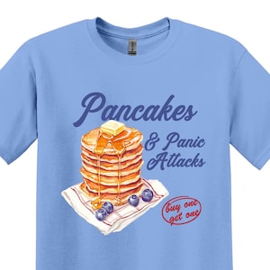 Könnte beinhalten: Ein hellblaues T-Shirt mit einem Grafikdruck von einem Stapel Pfannkuchen mit Butter und Sirup, Blaubeeren und dem Text "Pancakes & Panic Attacks" und "buy one get one".