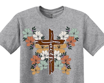 Könnte beinhalten: Ein heidefarbenes T-Shirt mit einem braunen Holzkreuz, das von bunten Blumen umgeben ist. Das Kreuz trägt die Aufschrift "Glaube".