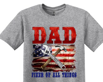 May include: A gray t-shirt with the text "DAD FIXER OF ALL THINGS" in red and blue. The text is overlaid on a distressed American flag design with a wrench and hammer.