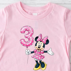 May include: A pink toddler t-shirt with a Minnie Mouse graphic holding a polka-dot number 3 balloon. The shirt is paired with blue jeans and pink canvas trainers. The Minnie Mouse graphic is wearing a pink dress and bow.