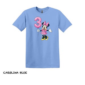 May include: Carolina blue t-shirt with a Minnie Mouse graphic holding a pink number 3 balloon. Minnie Mouse is wearing a pink polka dot dress and bow. The text "CAROLINA BLUE" is at the bottom of the image.