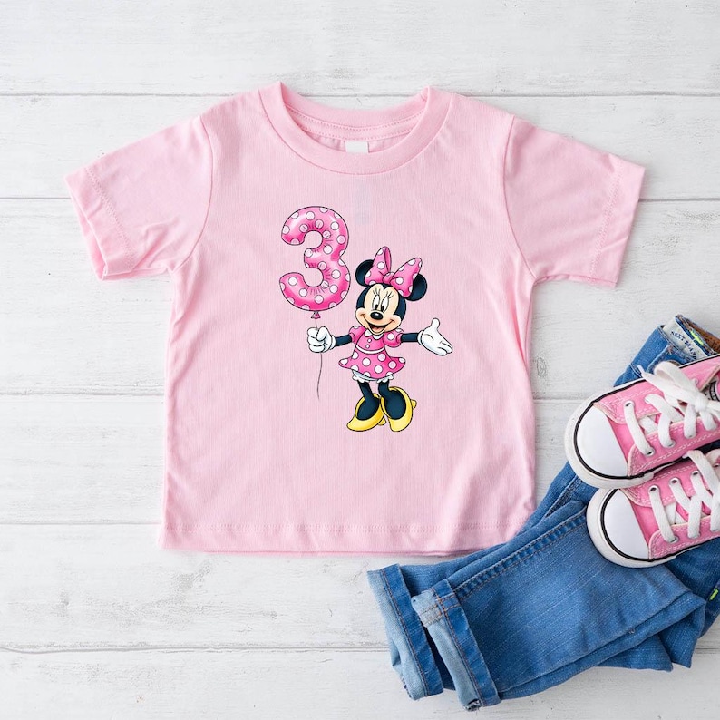 May include: A pink toddler t-shirt with a Minnie Mouse graphic holding a polka-dot number 3 balloon. The shirt is paired with blue jeans and pink canvas trainers. The Minnie Mouse graphic is wearing a pink dress and bow.