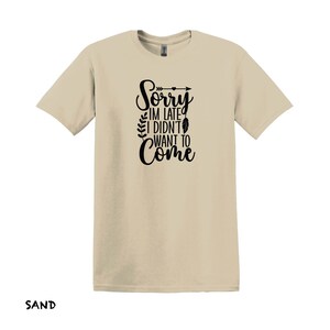 K&ouml;nnte beinhalten: Ein sandfarbenes T-Shirt mit einer schwarzen Grafik, auf der "Sorry, ich bin zu sp&auml;t, ich wollte nicht kommen" steht, mit einem kleinen Pfeil und einem Federdesign.