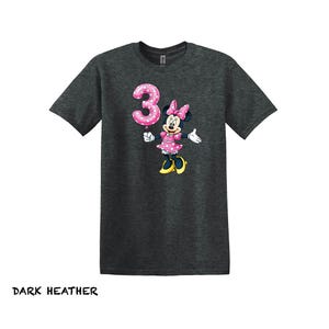 May include: Dark heather grey t-shirt with a Minnie Mouse graphic holding a pink number 3 balloon. Minnie Mouse is wearing a pink polka dot dress and bow. The text "DARK HEATHER" is at the bottom of the image.