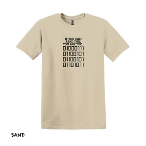 K&ouml;nnte beinhalten: Ein beiges T-Shirt mit einem schwarzen Aufdruck, der "IF YOU CAN READ THIS, YOU ARE TOO..." lautet, und einer Reihe von Bin&auml;rcode: "01000111 01100101 01100101 01101011".