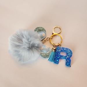 May include: A blue and white glitter keychain with a fluffy pom-pom, a tassel, and a blue letter R charm.