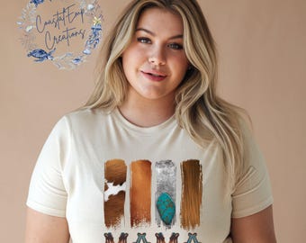 May include: A woman wearing a cream colored t-shirt with a graphic design of four vertical stripes in brown, tan, silver, and brown with a turquoise stone design. The text "MAMA" is printed below the stripes in a turquoise and brown design.
