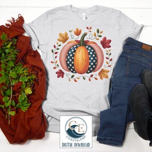 May include: A light gray t-shirt featuring a colorful pumpkin design. The pumpkin has orange, blue polka dot, and red gingham sections, surrounded by fall leaves and vines. The shirt is paired with blue jeans and black boots. The image includes a logo that says "Coastalemp Digital Download".