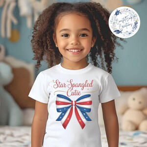 Puede incluir: Una camiseta blanca con un lazo rojo, blanco y azul y el texto "Star Spangled Cutie" en letras doradas.