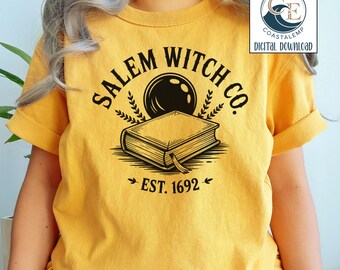Può includere: T-shirt giallo senape con grafica nera di un libro, una sfera di cristallo e la scritta "SALEM WITCH CO. EST. 1692". La maglietta ha maniche corte e una vestibilità rilassata. Un logo è nell'angolo in alto a destra.