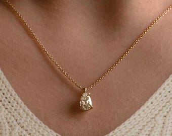 May include: A delicate gold necklace featuring a pear-shaped diamond pendant. The pendant is suspended from a fine chain, creating a simple yet elegant design. The necklace is worn against a cream-colored sweater.