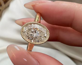 May include: A gold ring featuring an oval-cut diamond with a halo of smaller stones. The ring's band is a simple, polished gold. The diamond is the focal point, sparkling with numerous facets. The ring is held up, showcasing its design.