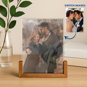 Puede incluir: Un marco de fotos de acrílico transparente con una base de madera muestra a una pareja abrazándose. El marco es rectangular y se mantiene en posición vertical. Una imagen más pequeña de una pareja está en la esquina superior derecha. Un jarrón de cristal con una planta verde está a la izquierda.