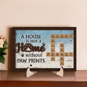 May include: A wooden sign with a blue and white background featuring the text "A house is not a home without paw prints". The text is surrounded by a brown frame and a scrabble tile design spelling out the names "Buddy", "Bailey", "Sunny", and "Peanut".
