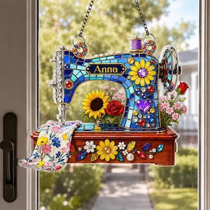 May include: A hanging ornament shaped like a sewing machine with a blue stained-glass pattern, the name Anna, and floral accents including sunflowers and roses. A piece of fabric with a floral print hangs from the needle area.