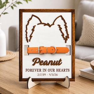 Puede incluir: Una placa conmemorativa rectangular de madera con marco marrón oscuro y centro blanco. Presenta un recorte marrón oscuro de orejas de perro, un collar de cuero naranja con hebilla metálica y el texto "Peanut" y "FOREVER IN OUR HEARTS 2/7/19 - 5/3/26".