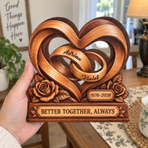 May include: A carved wooden heart sculpture featuring two interlocking rings engraved with the names Adrian and Violet. The base displays the text BETTER TOGETHER, ALWAYS and the date 1976-2026, accented with sculpted roses on each side.