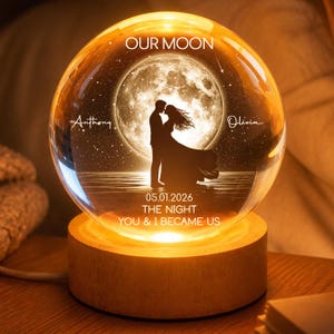 May include: A glowing glass sphere with a wooden base. Inside, a silhouette of a couple under a full moon is visible. The sphere is inscribed with the words "OUR MOON", names, and a date. The base emits a warm light.