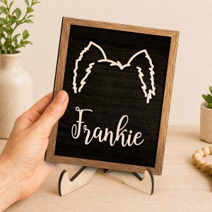 Puede incluir: Un letrero rectangular de madera con fondo de veta de madera negra, que presenta un recorte claro de orejas de perro en la parte superior y el nombre Frankie en letra cursiva debajo, todo enmarcado en un borde de madera marrón claro sobre un soporte.