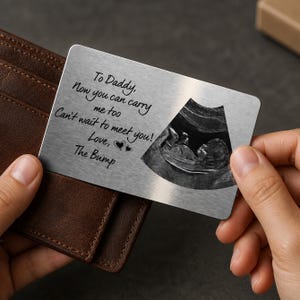 Może przedstawiać: Srebrna metalowa karta z napisem "To Daddy, Now you can carry me too, Can't wait to meet you! Love, The Bump." Obraz USG dziecka znajduje się po prawej stronie karty. Karta jest trzymana przed brązowym skórzanym portfelem.