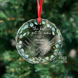 May include: A clear glass Christmas bauble with a green and gold floral design. The bauble features the text "the best is yet to come" and "ENGAGED -2024-" with the names Ethan and Sarah.