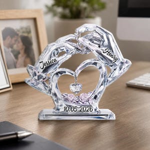 May include: A clear crystal sculpture featuring two hands forming a heart shape, with the names Jessica and Henry engraved on the wrists. A small heart pendant hangs in the center above two light pink roses, with the date 10.05.2026 printed on the base.