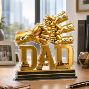 Könnte beinhalten: Ein goldfarbenes Dekorationsobjekt mit dem Wort "DAD" in großen Buchstaben. Über den Buchstaben befinden sich drei goldene Fäuste, jede mit einem Namen: Justin, Alex und Marley. Die Basis lautet "THE MAN THE MYTH THE LEGEND".