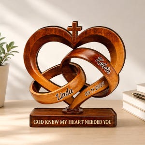 May include: A dark brown wooden heart sculpture featuring two interlocking loops and a small cross at the top. The piece is engraved with the names Linda and Kelvin, the date 03.11.2021, and the text GOD KNEW MY HEART NEEDED YOU on the base.