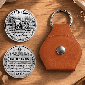 May include: Two circular silver-toned metal tokens with black engraved text and illustrations, alongside a brown leather keychain pouch with a silver snap button. The tokens feature mountain scenes and messages addressed to a son from a dad.