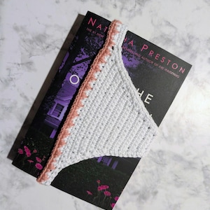 May include: A white crochet bookmark with a pink border rests on a book with the title "The Haunting" by Natasha Preston. The bookmark is triangular and handmade, perfect for marking pages.