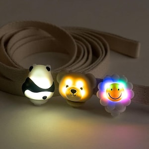 May include: Three light-up animal-shaped clips attached to a beige fabric strap. The clips are a panda, a bear, and a flower with a smiley face. The panda is white with black markings and glows white. The bear is brown and glows yellow. The flower is white with a yellow smiley face and glows in multiple colors.