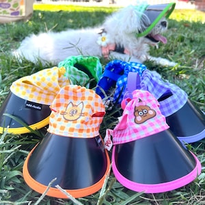 May include: Five colorful dog sun hats with different patterns and cartoon faces. The hats have a black visor and a colorful trim. The hats are on a green grassy surface.