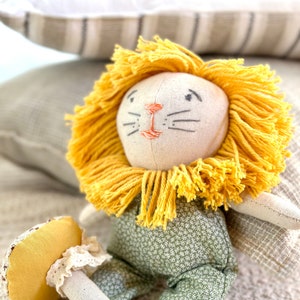 Puede incluir: Un juguete de peluche de león con una melena amarilla, que lleva pantalones verdes con flores blancas. El león tiene una cara blanca con ojos negros bordados y una nariz rosa.