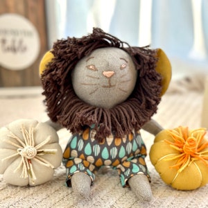Puede incluir: Un juguete de peluche de león gris con pelo marrón, que lleva un atuendo estampado en azul y marrón, sentado sobre una superficie de punto blanco con dos pequeñas calabazas de tela.