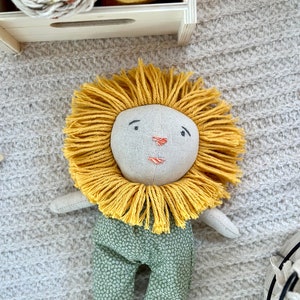 Puede incluir: Un juguete de peluche de león hecho a mano con una melena amarilla y pantalones de estampado verde. El león tiene una cara cosida con ojos negros y una nariz roja.
