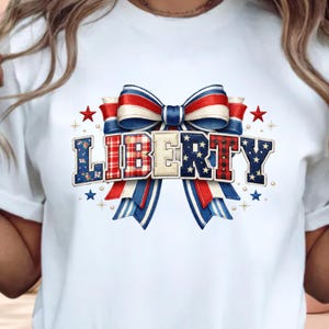 Puede incluir: Una camiseta blanca con un gráfico grande de un lazo rojo, blanco y azul atado detrás de la palabra LIBERTY. Las letras están rellenas con varios patrones, incluyendo flores, cuadros y estrellas, rodeadas de pequeñas estrellas y puntos.