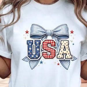 May include: A light blue t-shirt featuring a graphic of a large blue and white striped bow centred above the letters USA. The U is blue with white flowers, the S is red and white gingham, and the A is cream with blue stars and small red stars.