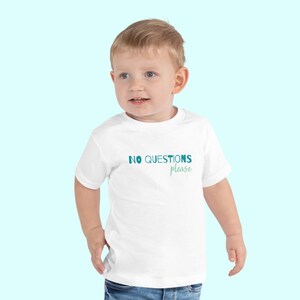 Puede incluir: Una camiseta blanca para niño con el texto "NO QUESTIONS please" en verde azulado y verde azulado claro. El niño lleva vaqueros azules y está de pie frente a un fondo azul claro.