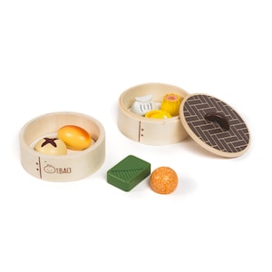 May include: Wooden toy dim sum set with two bamboo steamers and various food items. The set includes a green rectangular piece, an orange ball, and several yellow and white dim sum pieces. The steamer lids have a brown geometric pattern.