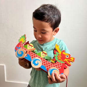 May include: A child plays with a wooden toy dragon boat with colorful characters. The boat has red, blue, and green details and wheels. The child is holding the toy in their hands.