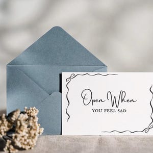 Puede incluir: Un sobre azul y una tarjeta blanca con el texto "Open When You Feel Sad" en escritura elegante. La tarjeta tiene detalles decorativos de borde negro. También hay un pequeño ramo de flores secas.