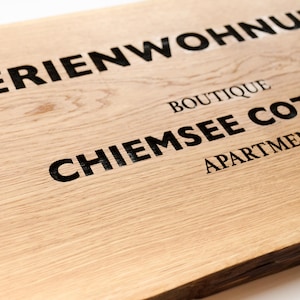 Könnte beinhalten: Ein rechteckiges, hellbraunes Holzschild mit sichtbarer natürlicher Maserung. Das Schild trägt eine schwarze Gravur mit dem Text FERIENWOHNUNG, BOUTIQUE, CHIEMSEE COTTAGE und APARTMENT. Die Kanten des Holzbretts sind dunkel und strukturiert.