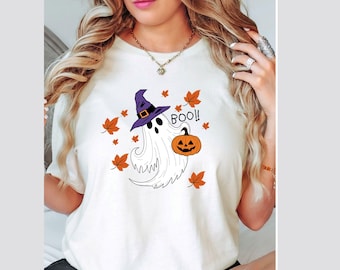 May include: A cream-colored t-shirt featuring a graphic of a white ghost wearing a purple witch hat and holding an orange jack-o'-lantern. The design includes the text "BOO!!" and several scattered orange and brown autumn leaves.